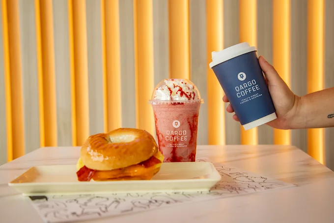 A breakfast spread featuring a bagel sandwich with bacon and cheese on a plate, alongside a frozen strawberry drink topped with whipped cream and a hot beverage in a cup.