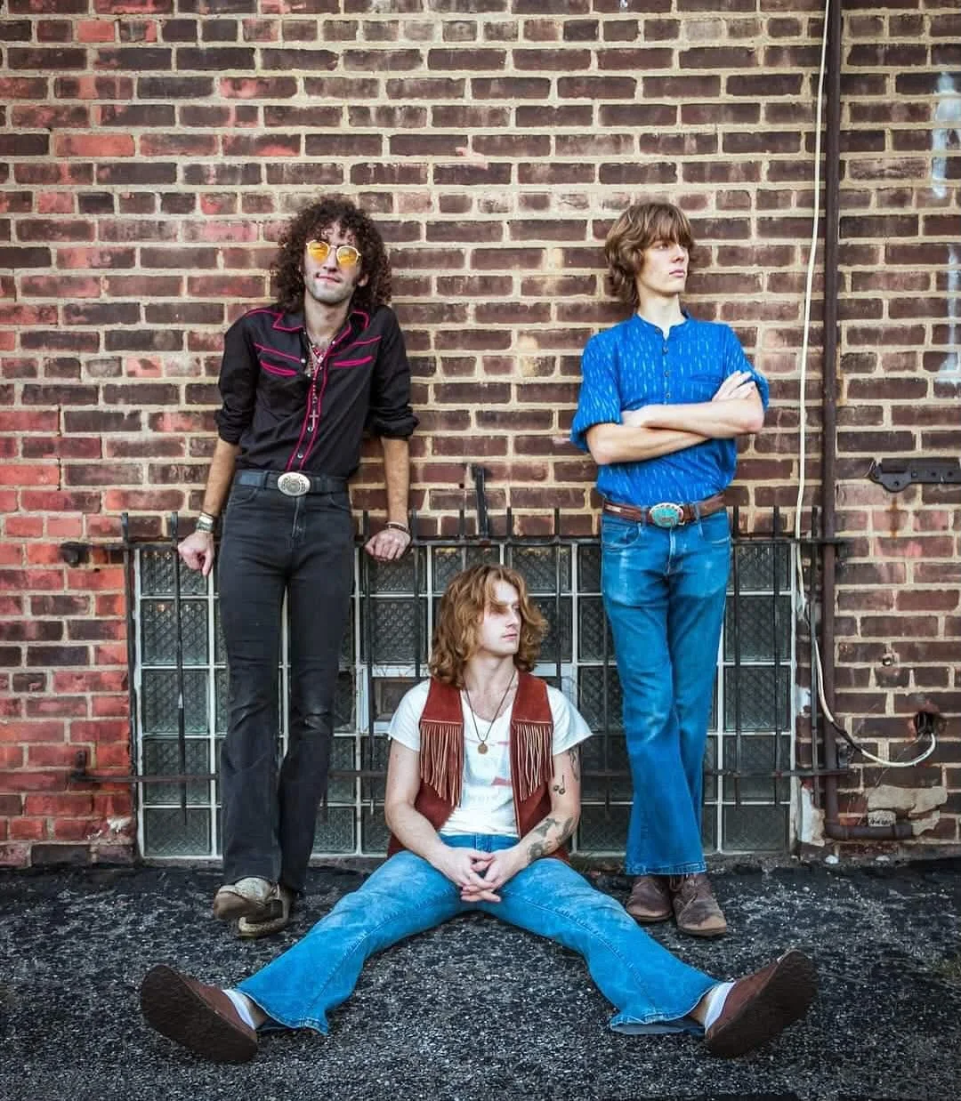 Three musicians posing against a brick wall in retro outfits
