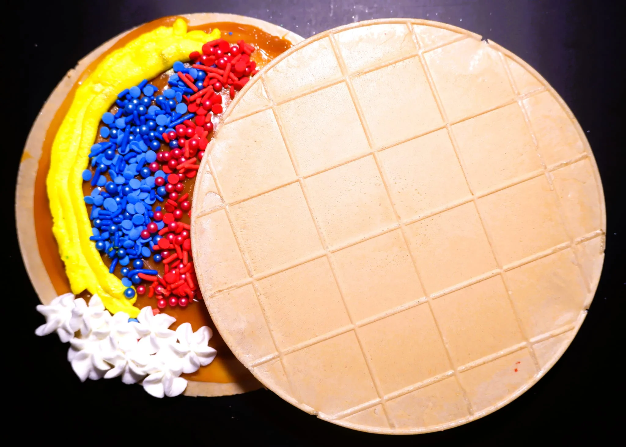 A decorated cake with colorful icing and sprinkles, partially covered by a cake lid.