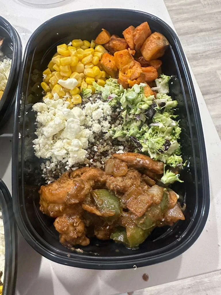 A lunch box containing corn, roasted sweet potatoes, a salad with broccoli, quinoa, and a portion of chicken in a savory sauce.