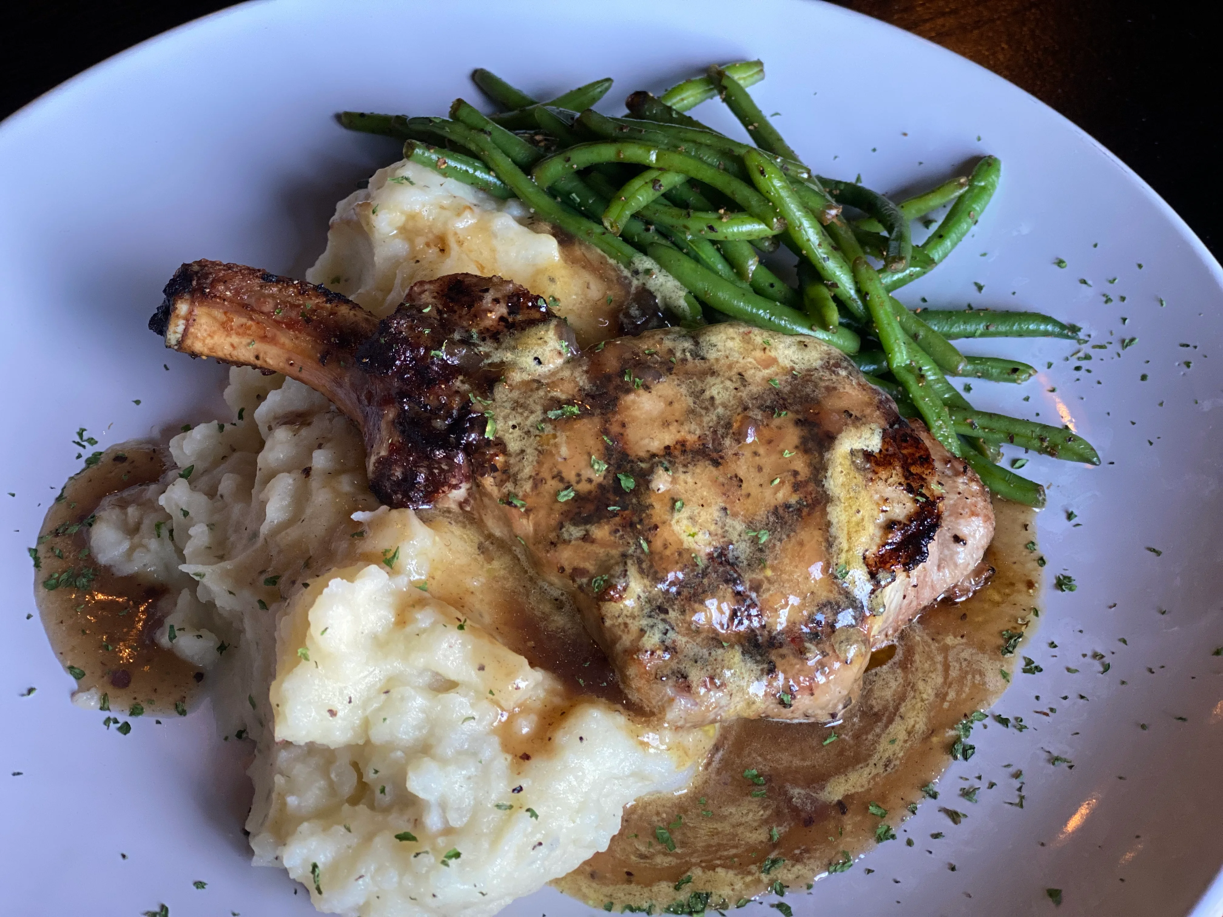A hearty dish featuring a grilled pork chop served over creamy mashed potatoes, accompanied by green beans and a savory sauce.