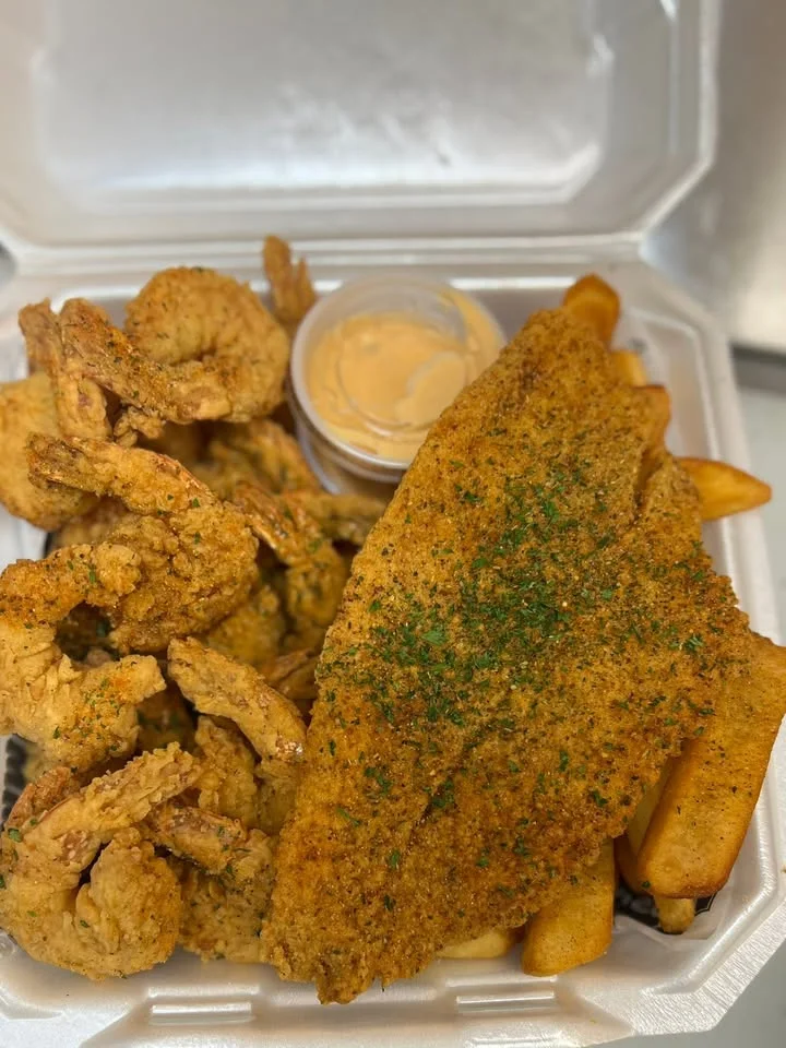 A takeout container featuring crispy fried shrimp, a seasoned fish fillet, French fries, and a small dipping sauce.