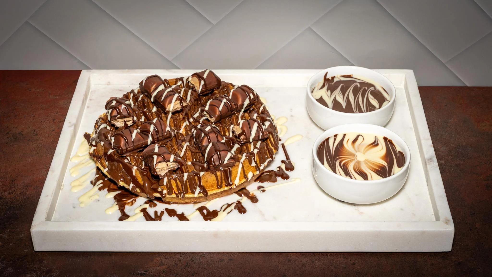 A decadent dessert featuring a round cake topped with chocolate drizzle and pieces, served with two bowls of chocolate sauce.