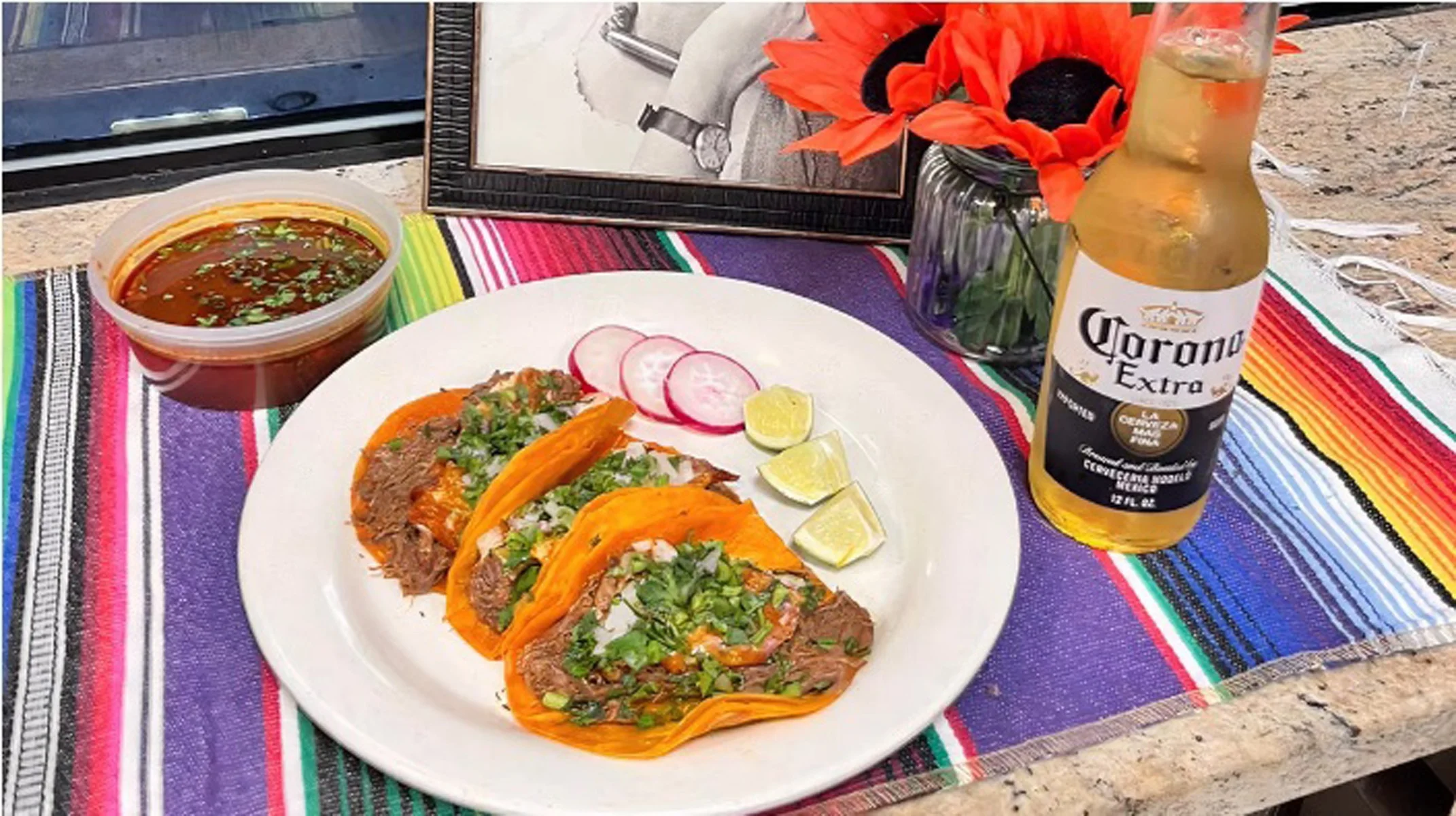 A serving of tacos with toppings such as meat, cilantro, and onions, accompanied by lime wedges and radishes. A bowl of salsa and a bottle of beer are on the side.