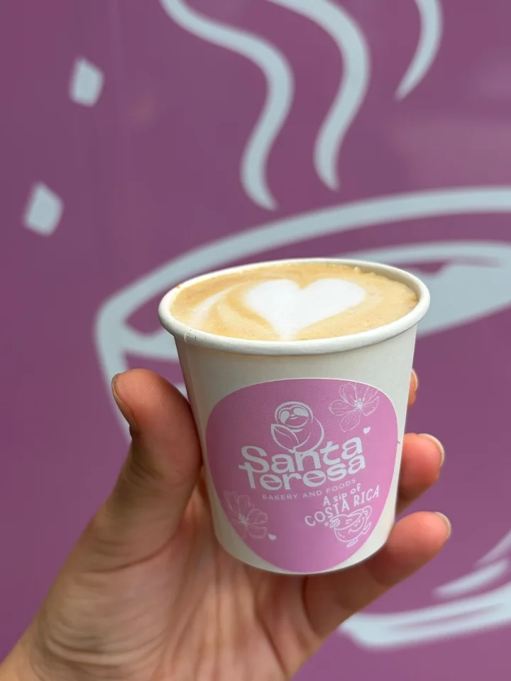 A small cup of latte with a heart design in foam, held by a hand. The cup features a pink design, set against a pink backdrop.