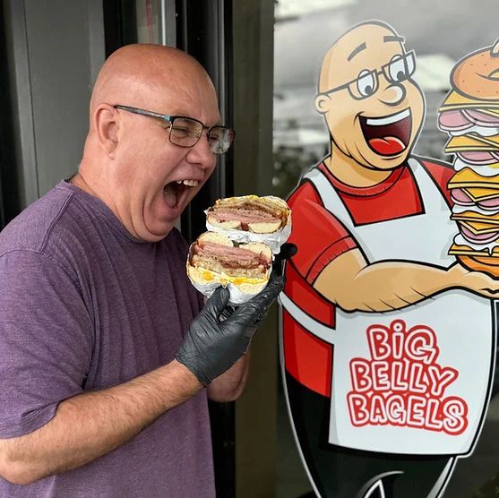 Excited chef holding a giant bagel sandwich outside Big Belly Bagels