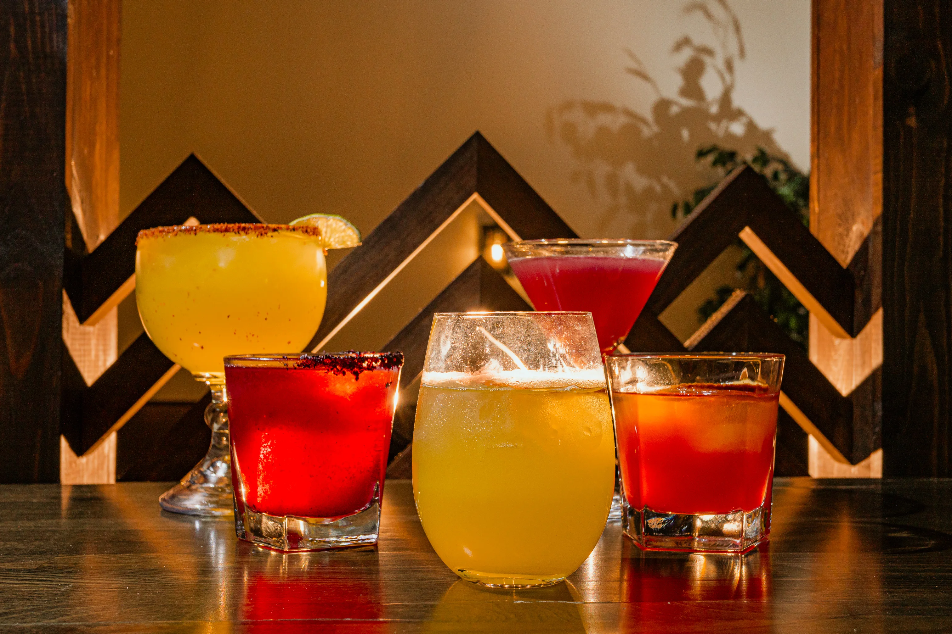 A variety of colorful cocktails arranged on a wooden table. The drinks include margaritas, a red cocktail with a sugared rim, and other vibrant beverages under warm lighting.