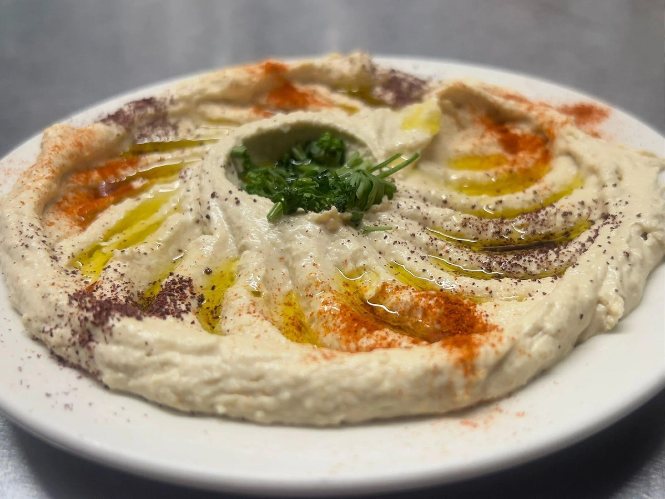 A plate of hummus garnished with olive oil, spices, and fresh herbs, showcasing a creamy texture with decorative swirls.