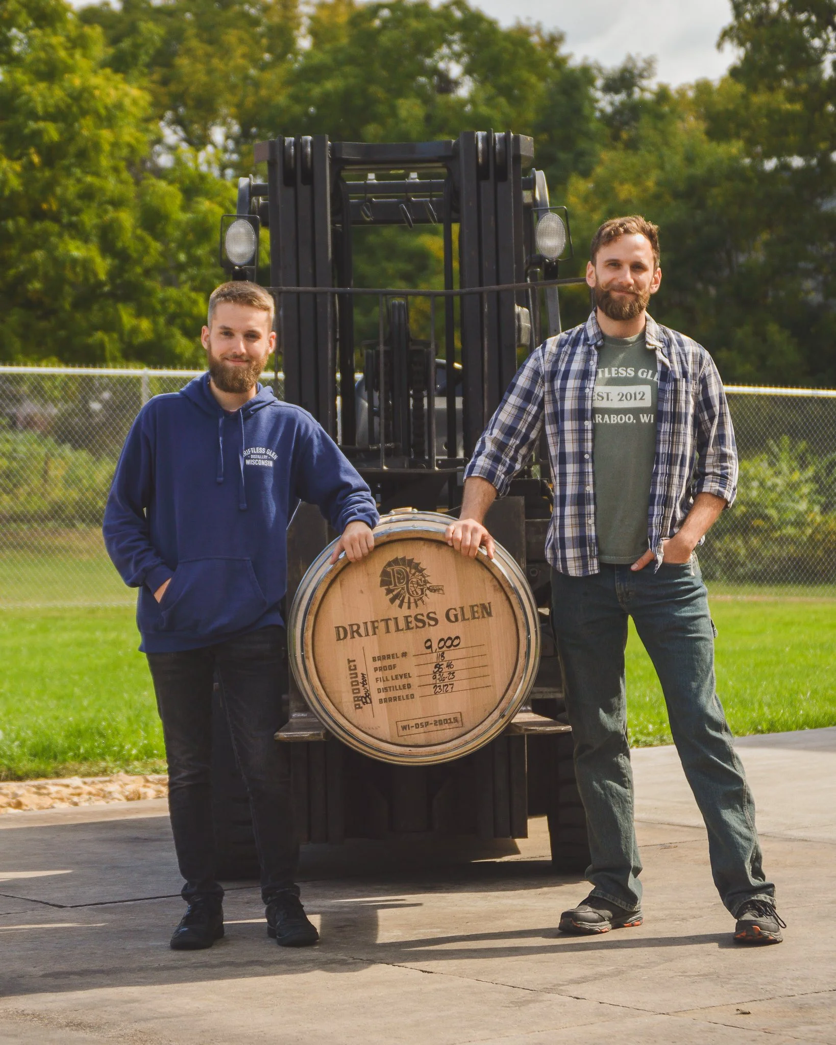 Two men stand with their hands resting on barrel #9000 outside on a forklift at Driftless Glen
