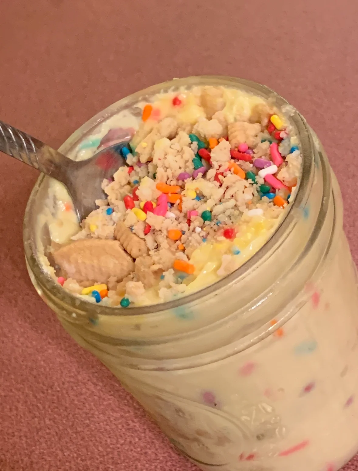 A mason jar filled with a creamy dessert topped with colorful sprinkles and crumbled cookies. A spoon rests beside the jar, ready for serving.