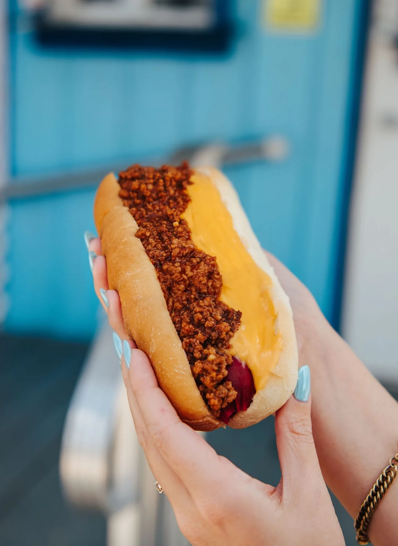 A hot dog with chili and melted cheese served in a soft bun. It is held by a hand against a colorful background.