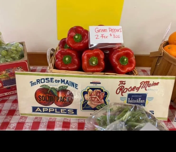 A basket of red bell peppers displayed with a price sign for green peppers. The background features different types of fruits and vegetables.