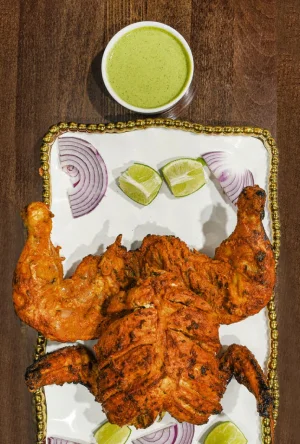 A whole roasted chicken, marinated and grilled, presented with lime wedges and onion slices, accompanied by a green dipping sauce.
