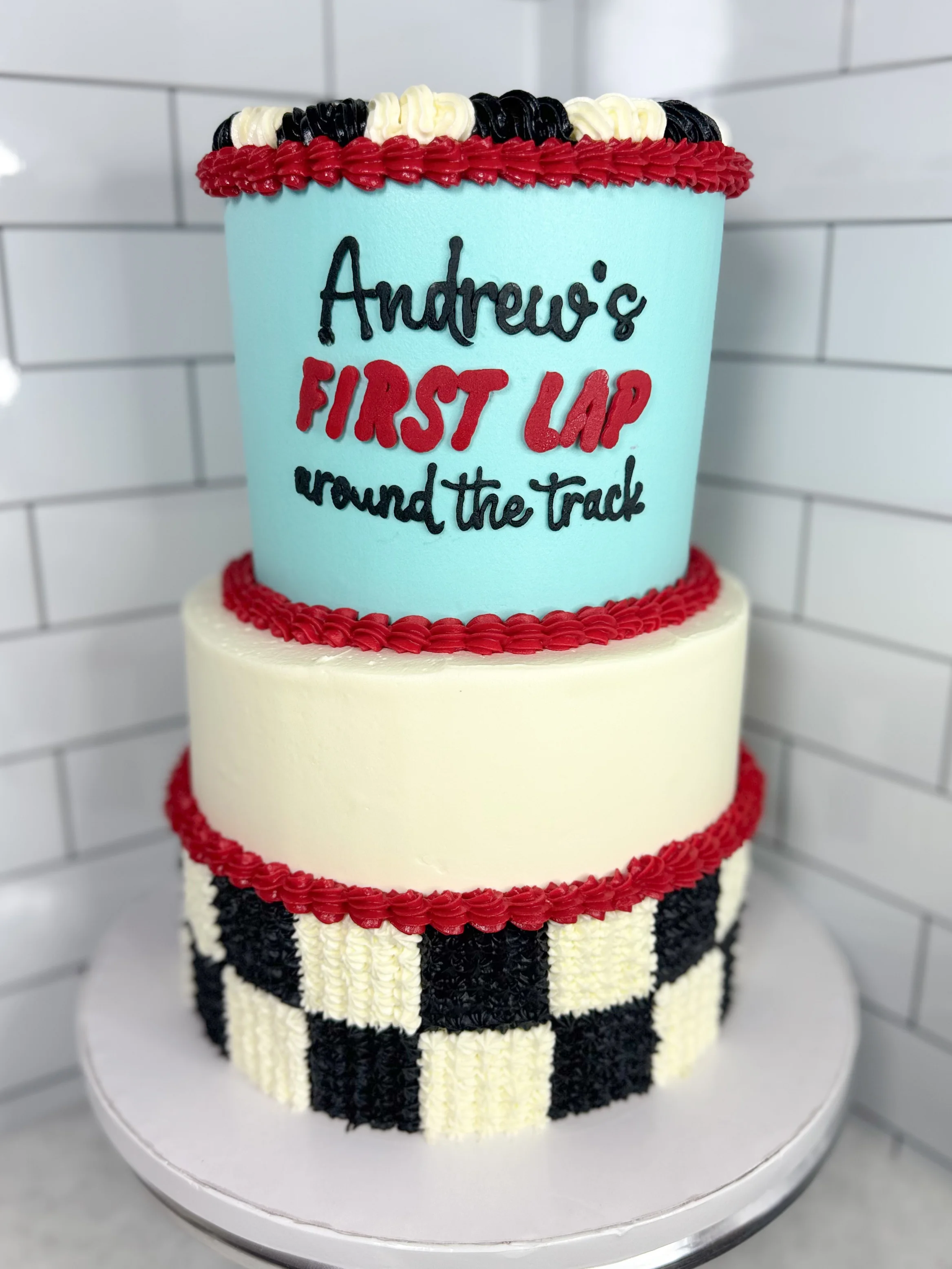 A race-themed two-tier cake decorated with a checkered pattern and festive message about a first lap.