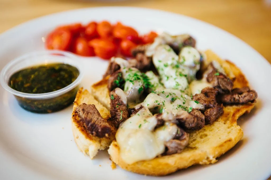 A savory dish featuring grilled meat topped with melted cheese and herbs on a piece of bread, accompanied by a side of cherry tomatoes and a green sauce.