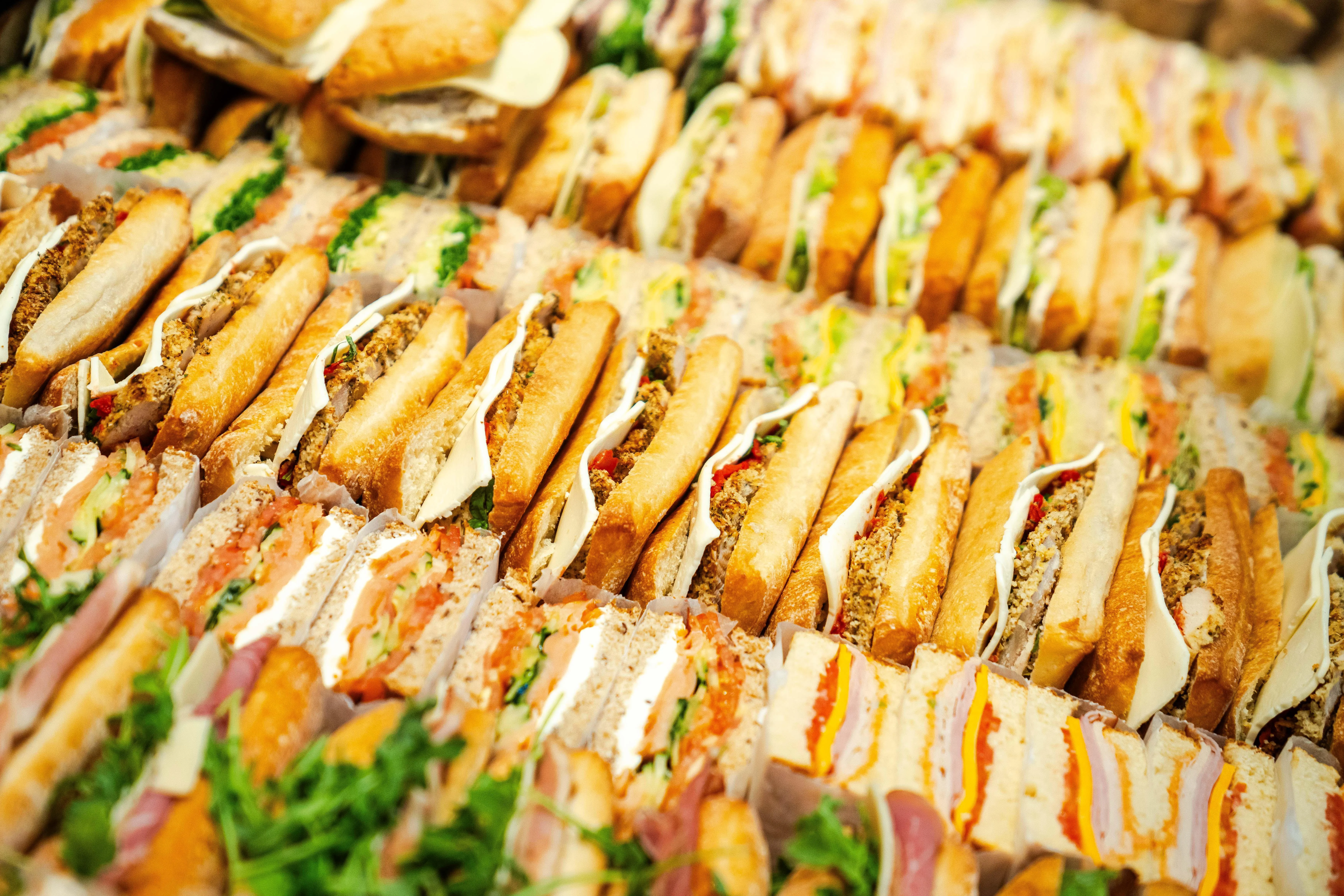 A large selection of sandwiches laid out in rows, featuring various meats, cheeses, and fresh vegetables.