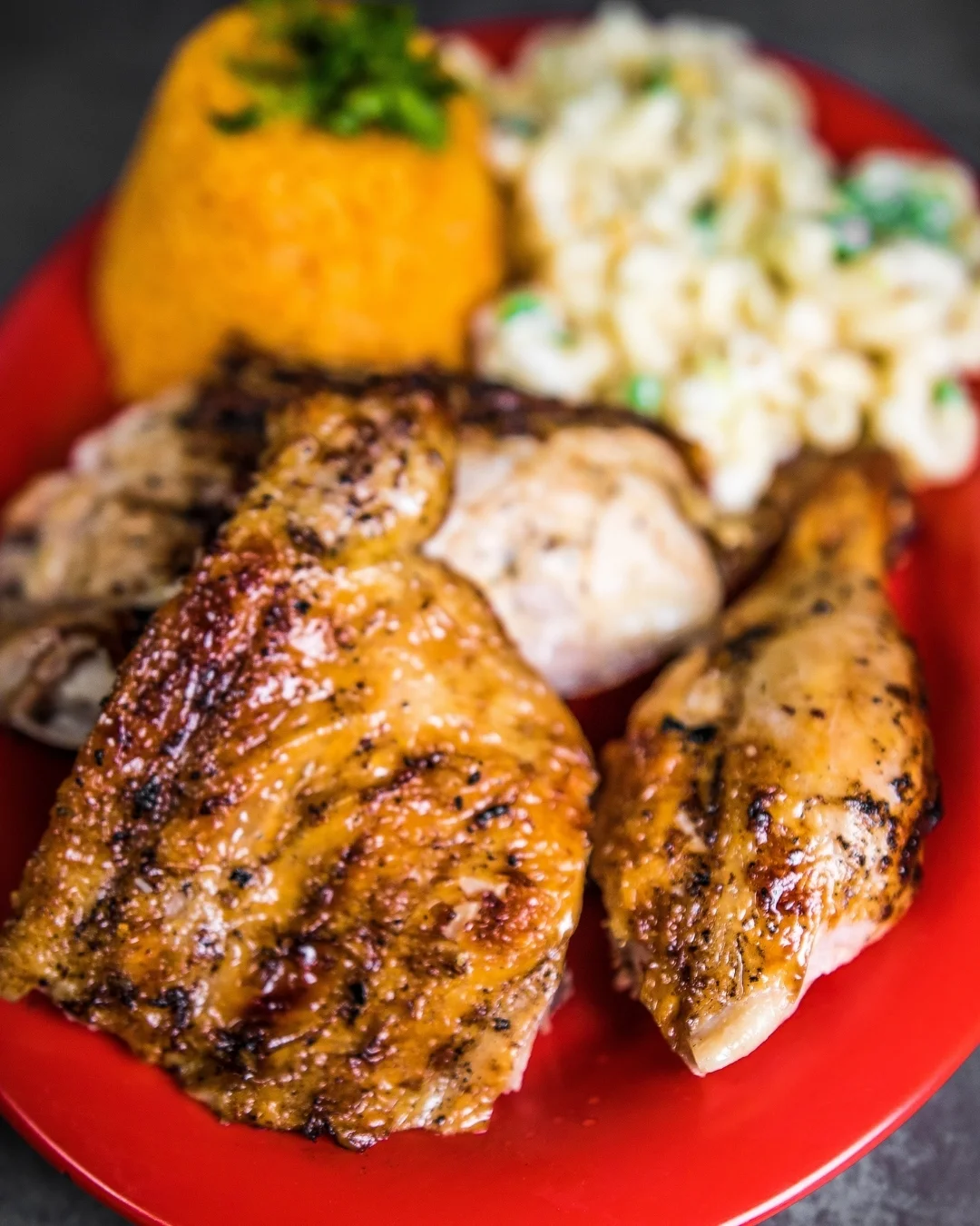 A serving of grilled chicken accompanied by a baked potato topped with cheese and a bowl of beans. The meal is presented on a red plate.