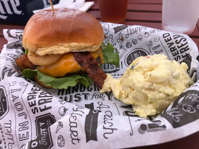 A juicy chicken burger with bacon, cheese, lettuce, and a side of creamy potato salad.