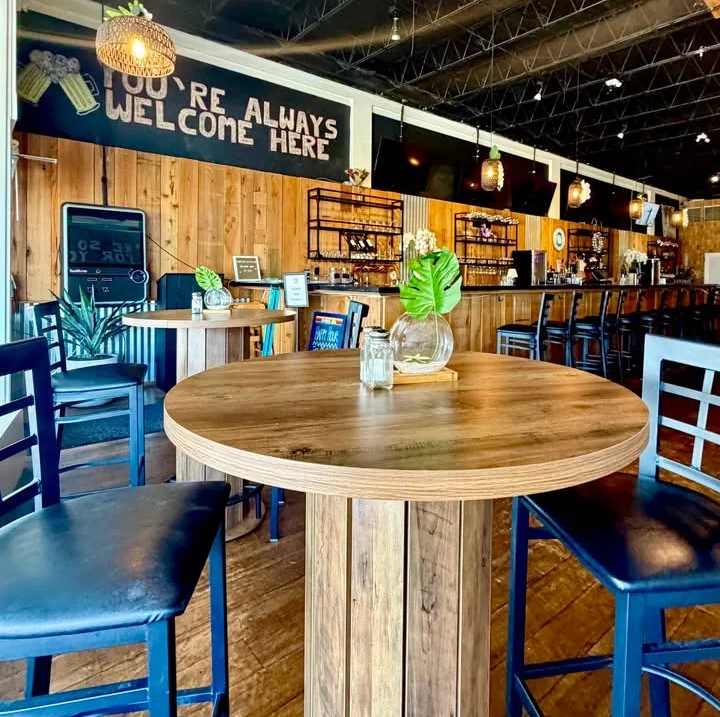 A view of a restaurant interior featuring a round wooden table and bar stools. The décor is welcoming, with a sign that reads 'YOU'RE ALWAYS WELCOME HERE'.