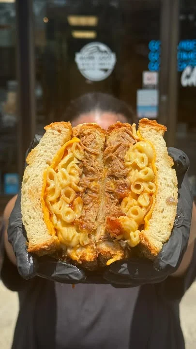 A close-up of a sandwich filled with pulled meat and macaroni and cheese, held by a person wearing black gloves. The sandwich is cut in half to reveal the filling.