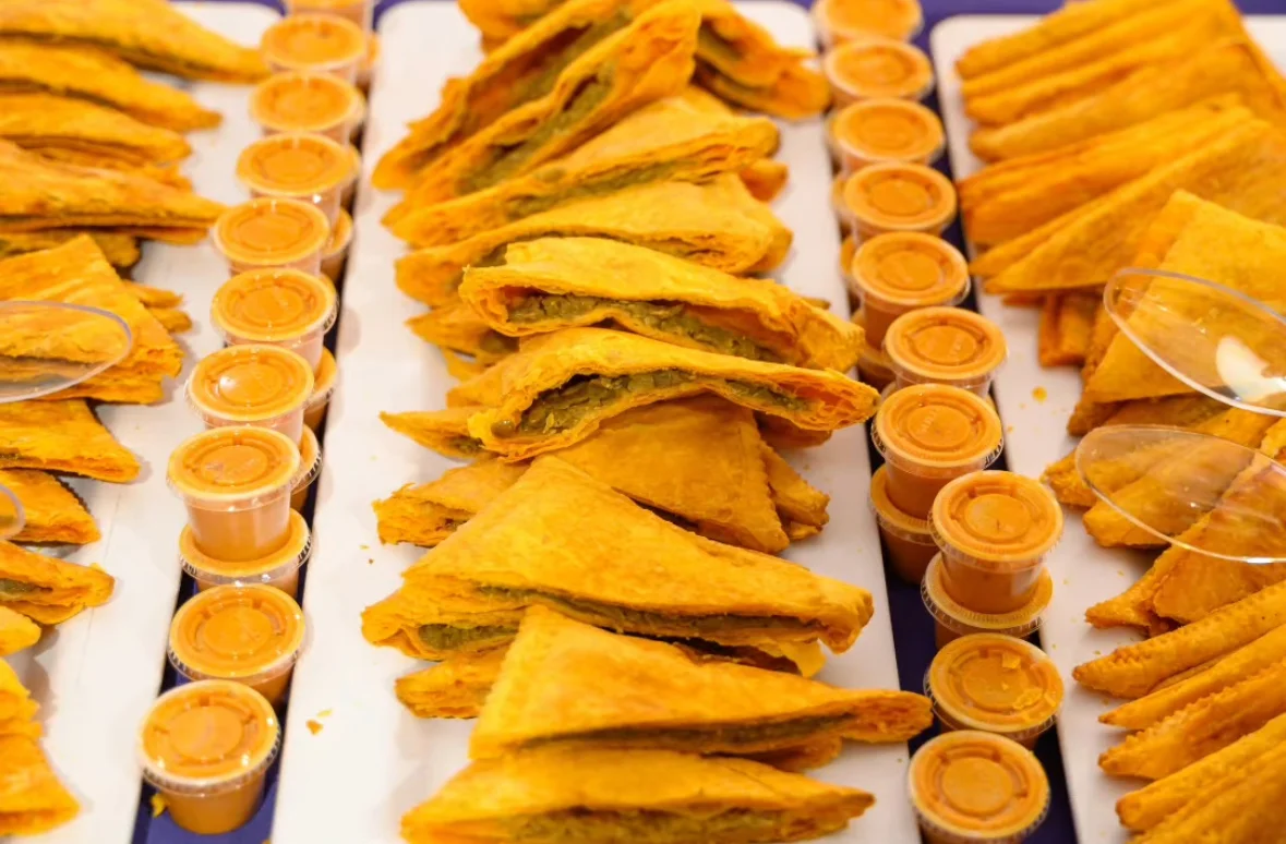 Golden triangular savory dish arranged in a row on a white tray, flanked by small cups of orange sauce, creating a warm and appetizing display.