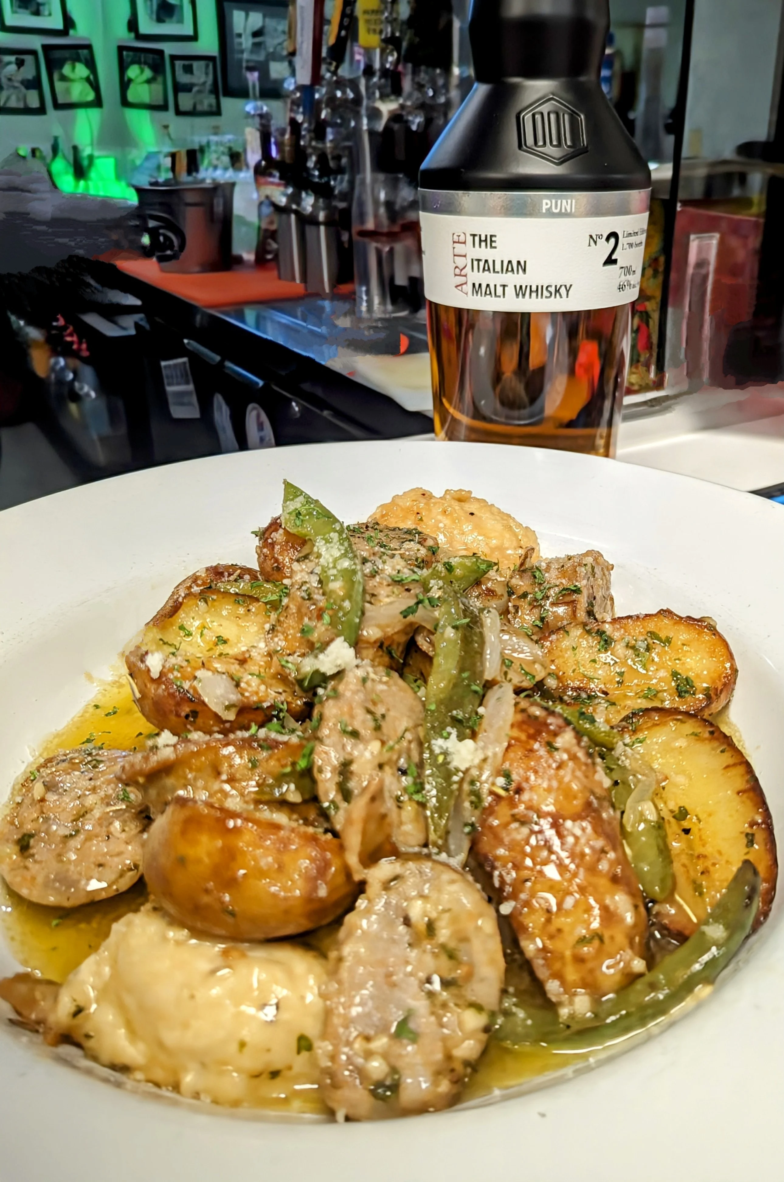 A savory dish featuring sliced sausage and potatoes tossed with green peppers and herbs on a white plate, alongside a bottle of whisky.