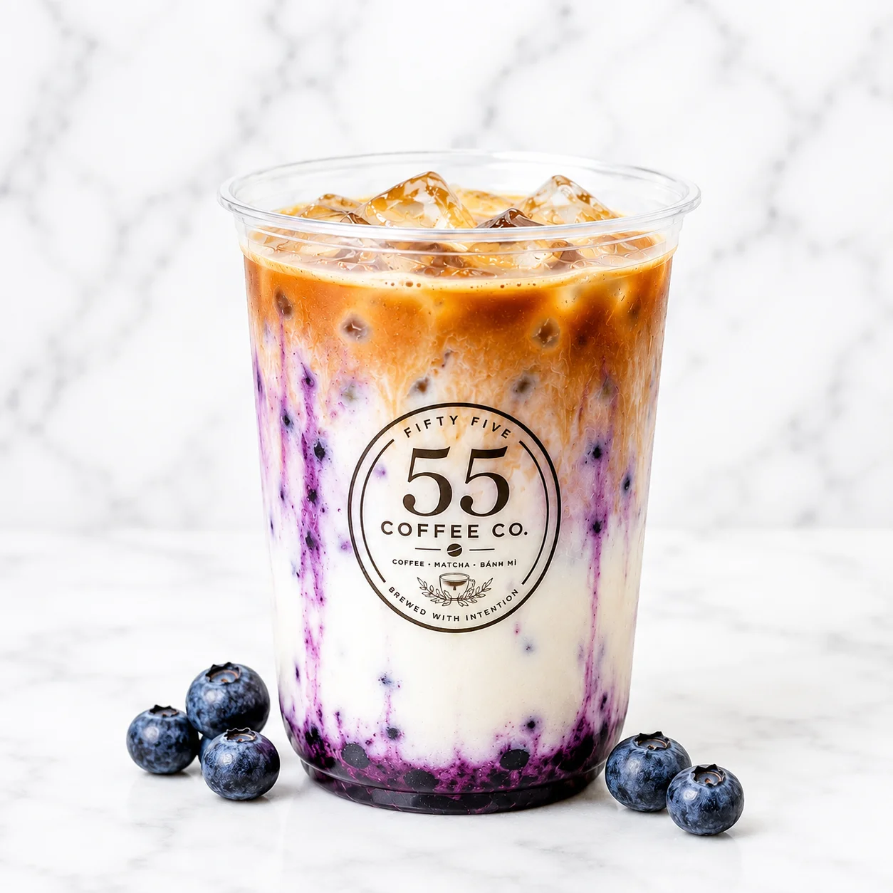 A glass cup filled with iced coffee, swirling with cream as it is being poured. The drink has a rich brown color and is served with ice.