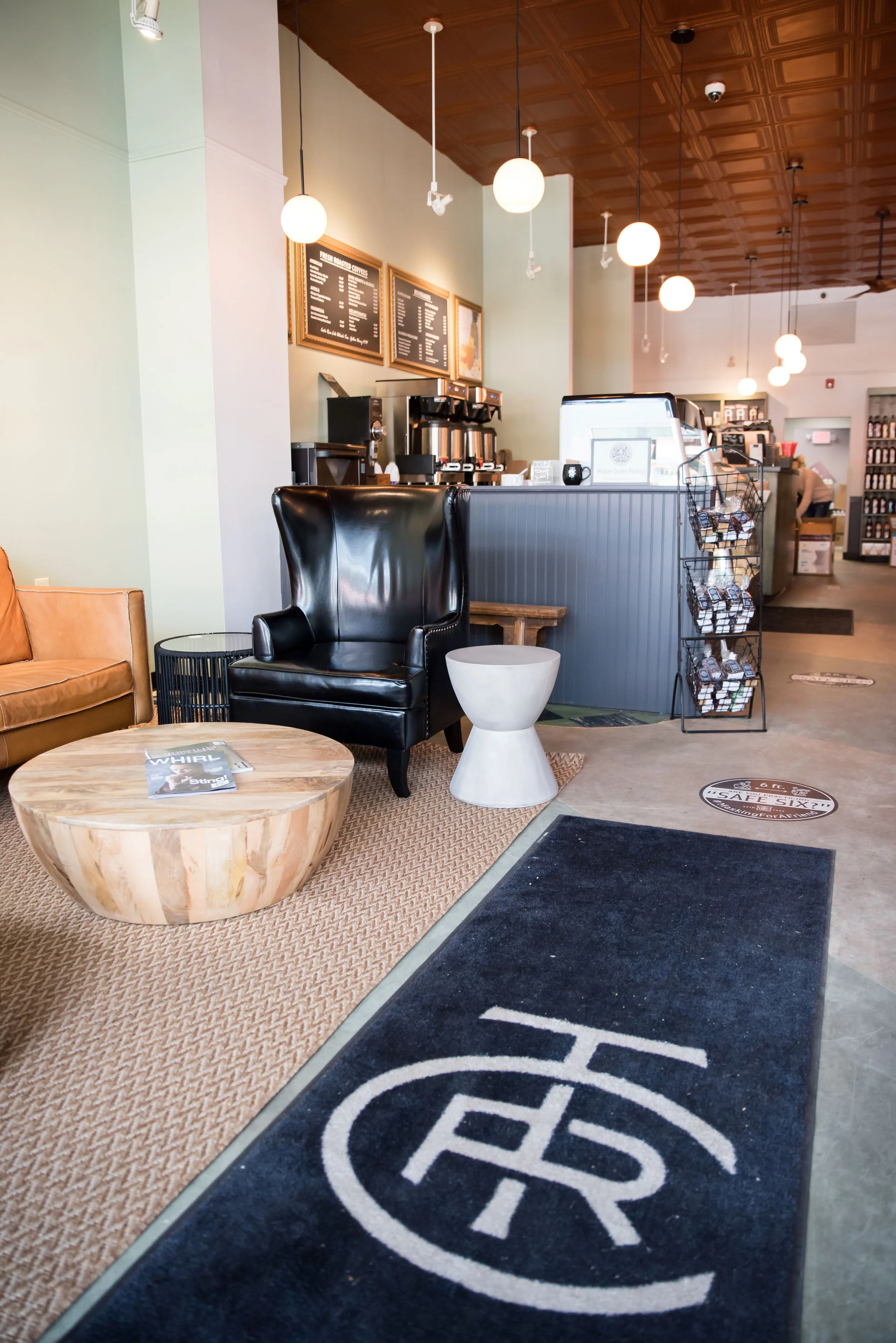 Chic coffee shop interior with leather chairs, round wooden table, and hanging globe lights. Coffee counter in background; welcoming, cozy atmosphere.