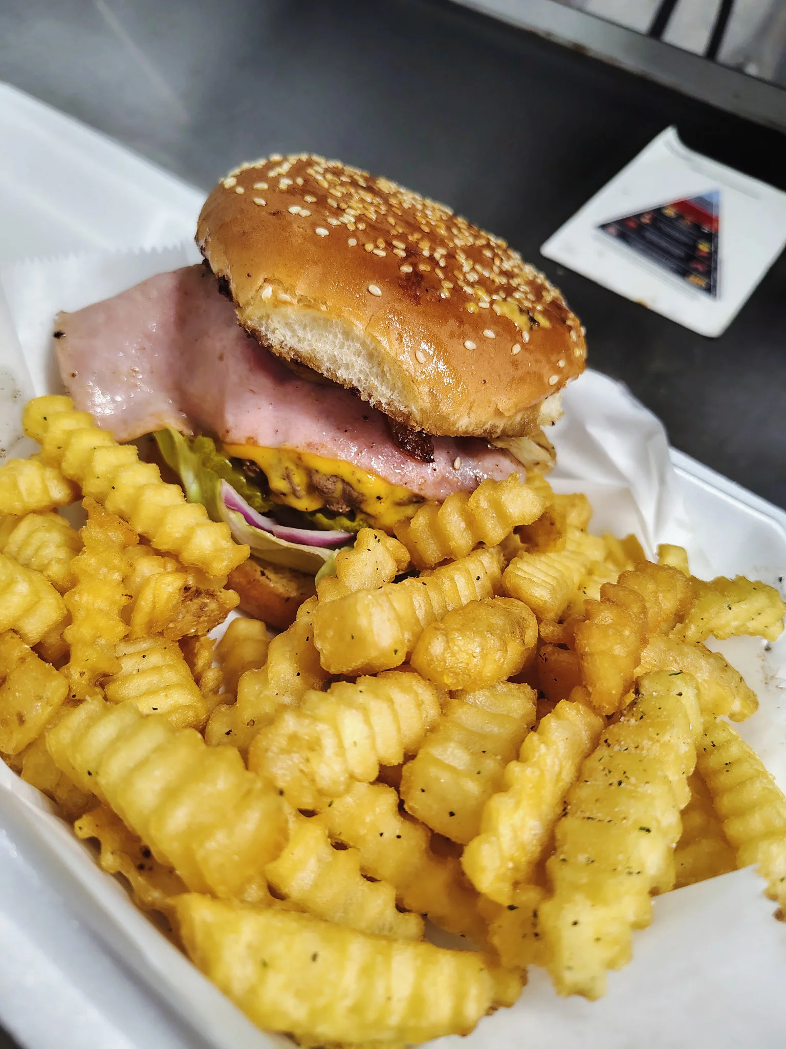 A close-up of a sesame seed bun burger alongside a generous serving of crinkle-cut fries. The burger is stacked with ham, cheese, and fresh vegetables.