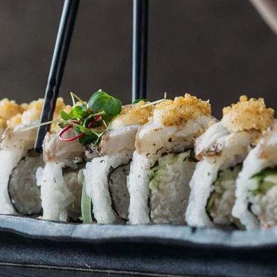 A platter of sushi rolls topped with crispy garnishes and delicate herbs, held with chopsticks.