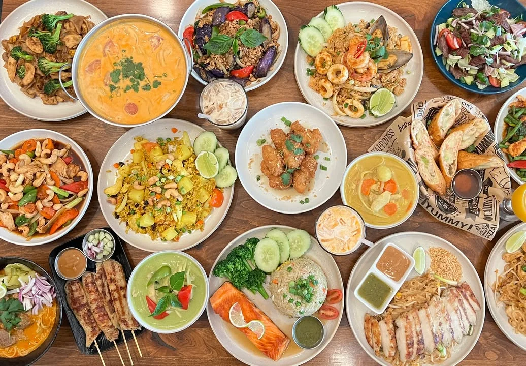 A variety of colorful dishes arranged on a wooden table, including stir-fried vegetables, curries, rice, and appetizers.