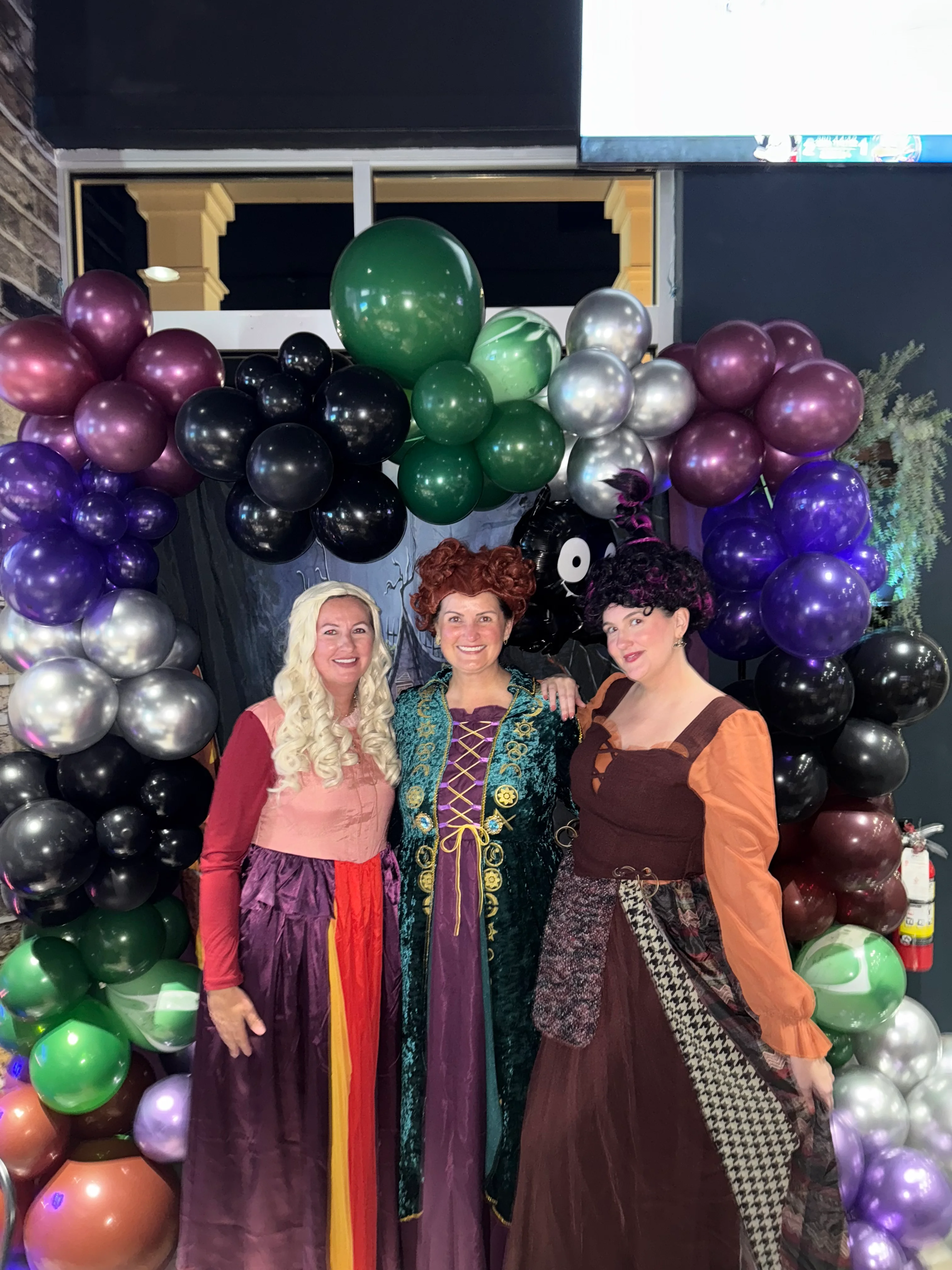 Three women in Halloween costumes posing in front of a balloon arch