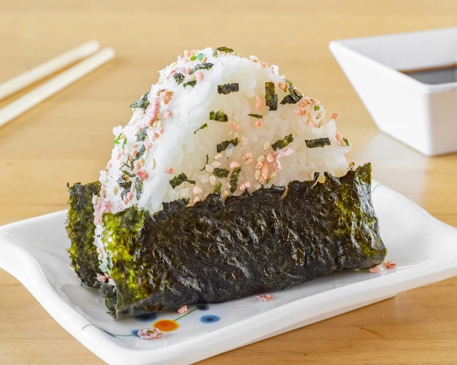 A rice ball wrapped in seaweed, sprinkled with seasoning and garnished with bits of dried fish, served with a small dish of soy sauce.