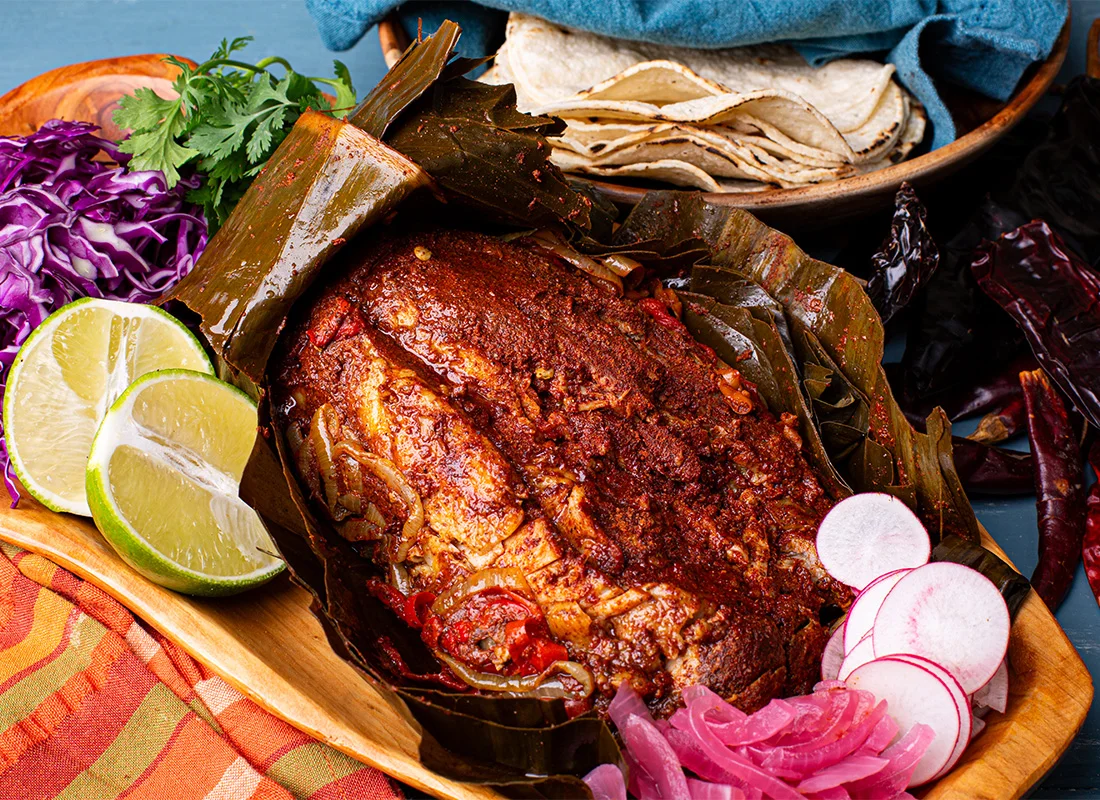 A traditional dish wrapped in banana leaves with colorful sides of lime, cabbage, and pickled onions.