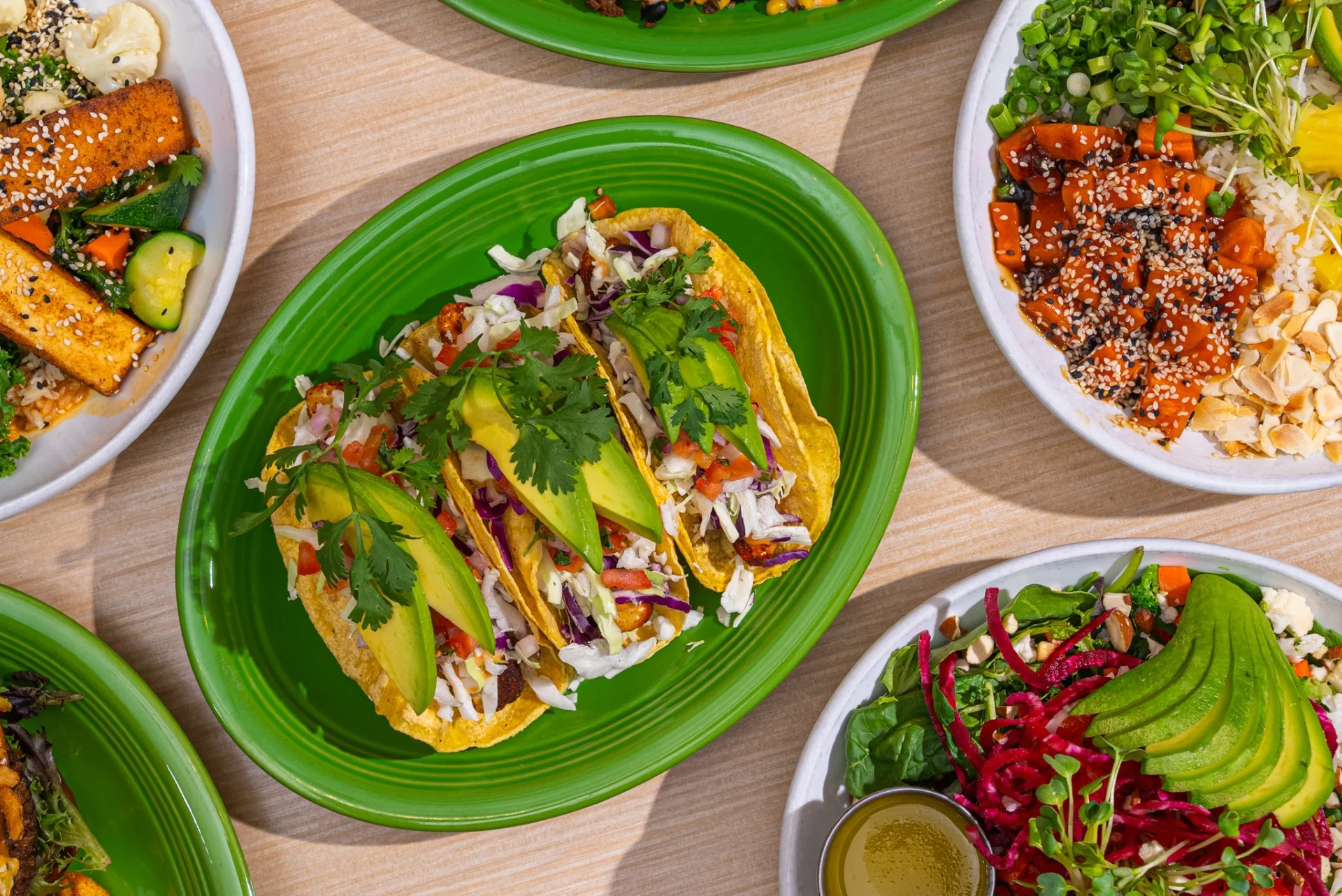 A colorful platter featuring three tacos filled with vegetables and topped with cilantro and avocado, accompanied by several small side dishes.