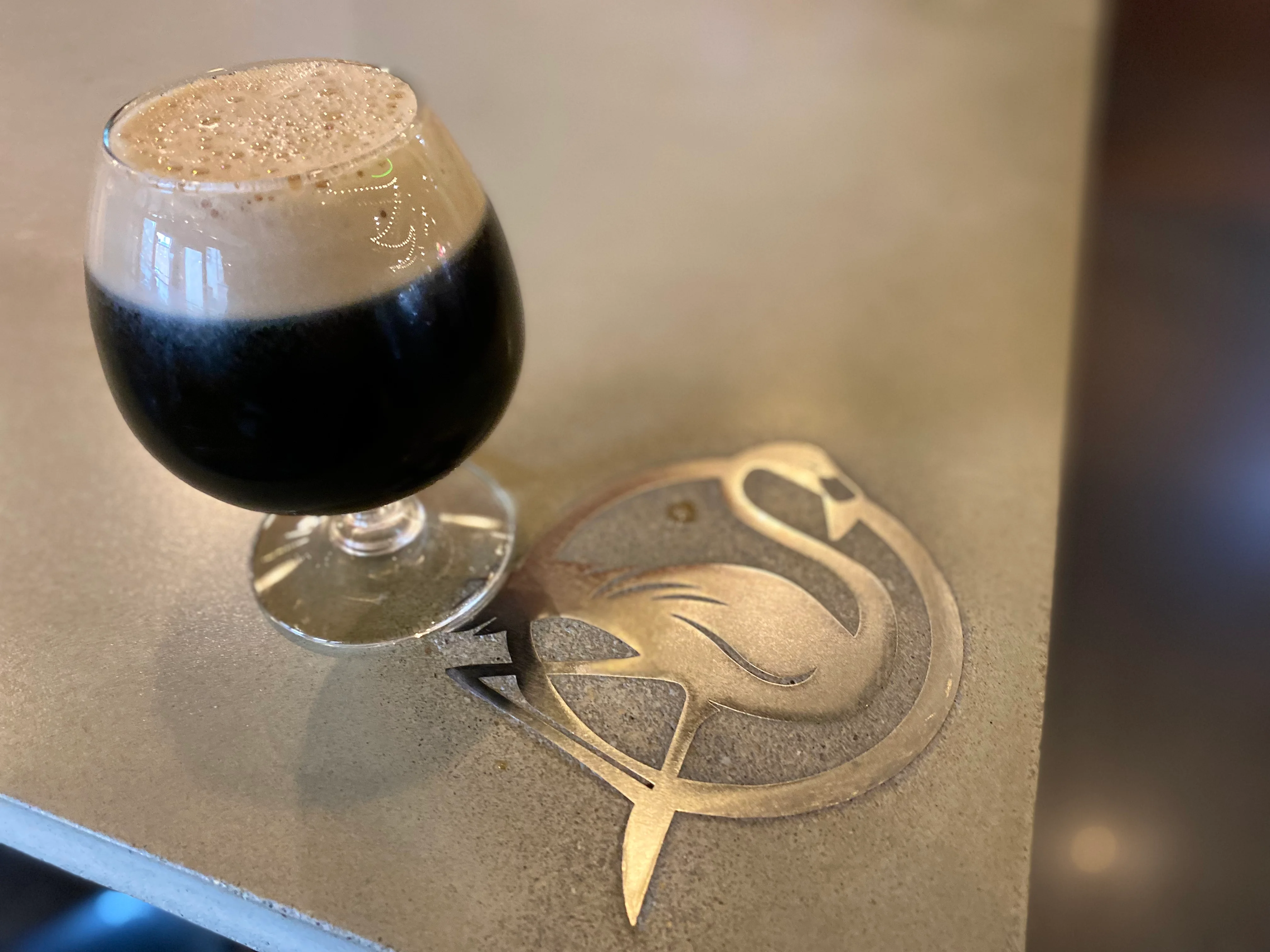 A snifter glass filled with a rich, dark stout at Isaac's Craft Kitchen & Brewery, topped with a creamy tan head, resting on a sleek bar counter featuring a metallic flamingo inlay.