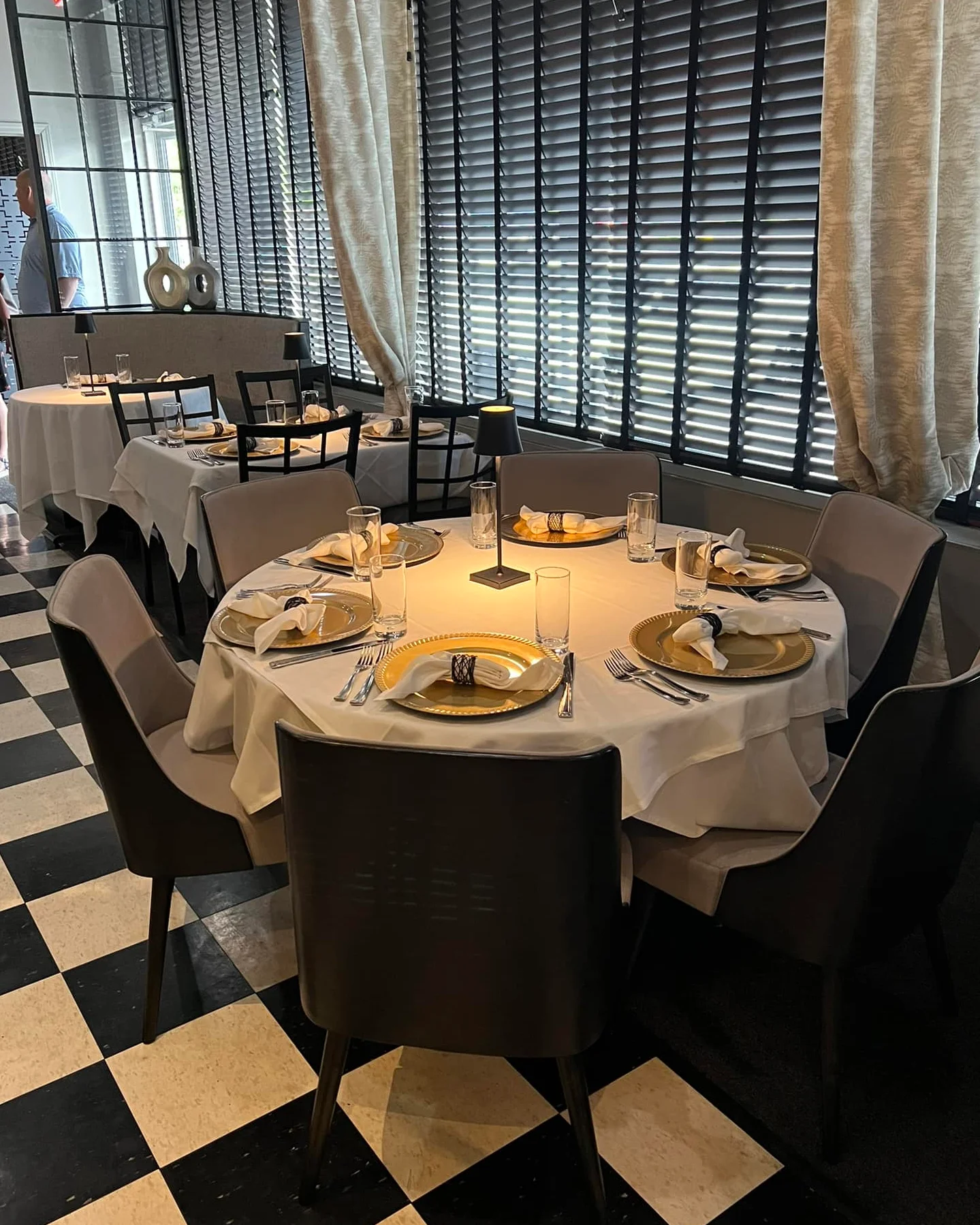 Chic dining space with a round table set for six, featuring gold charger plates, folded napkins in black rings, and tall water glasses. Black and white checkered floors and soft beige drapes add a classic touch, while black chairs and dim table lamps create a modern, elegant ambiance