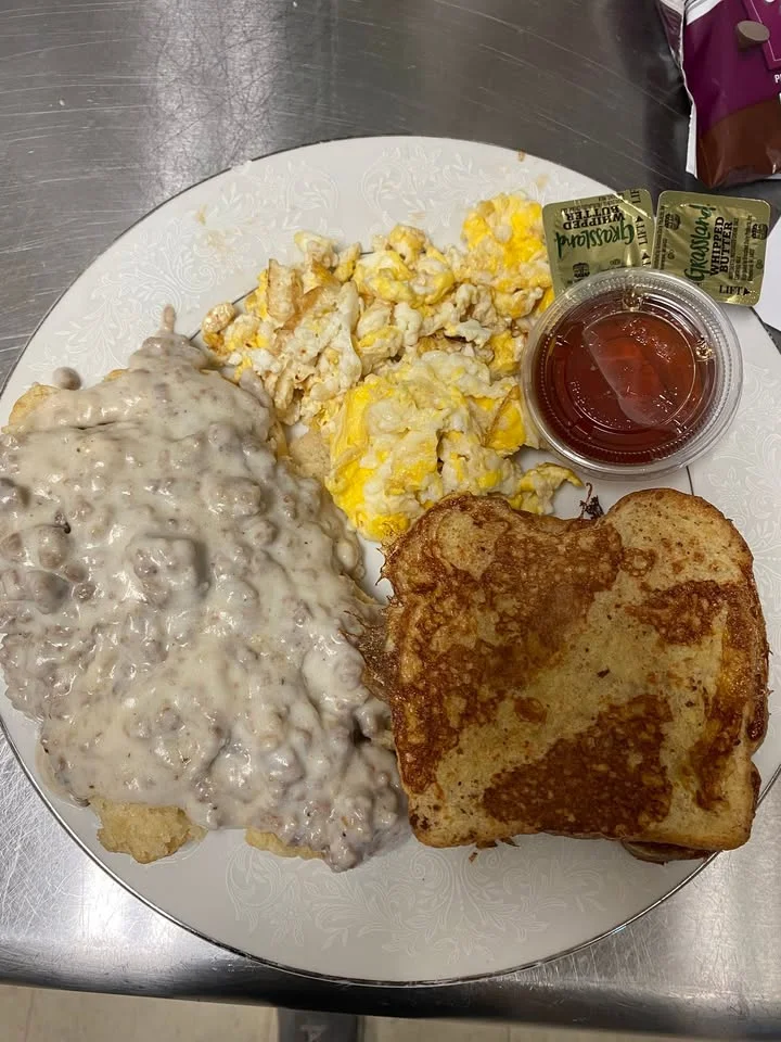 A plate featuring biscuits topped with creamy sausage gravy, scrambled eggs, and a slice of toast. A small side of salsa is also included.