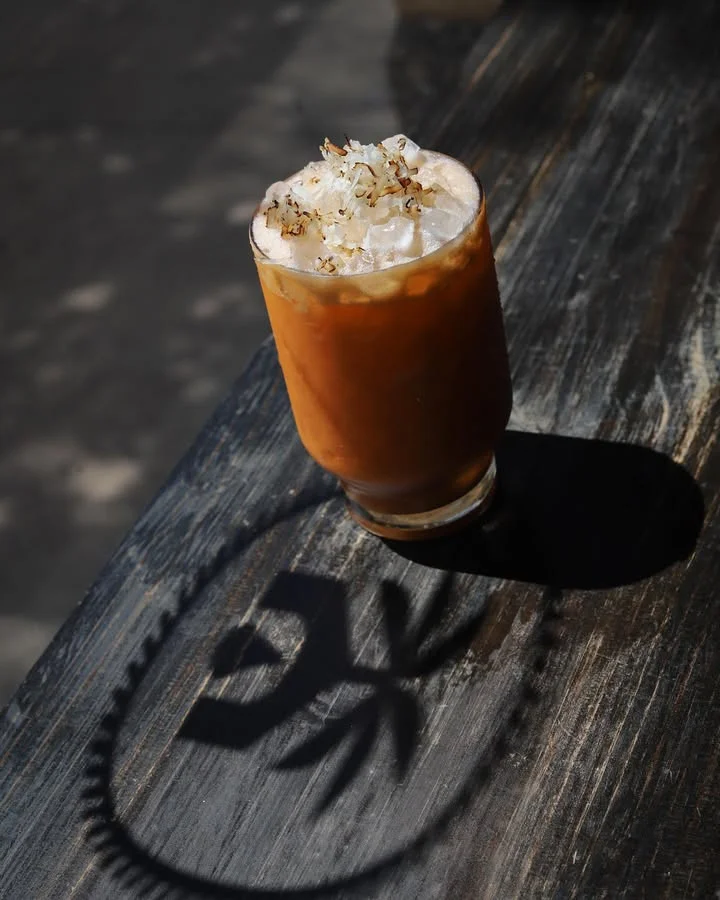 A glass of iced drink topped with toasted coconut flakes, casting a shadow on a wooden surface.