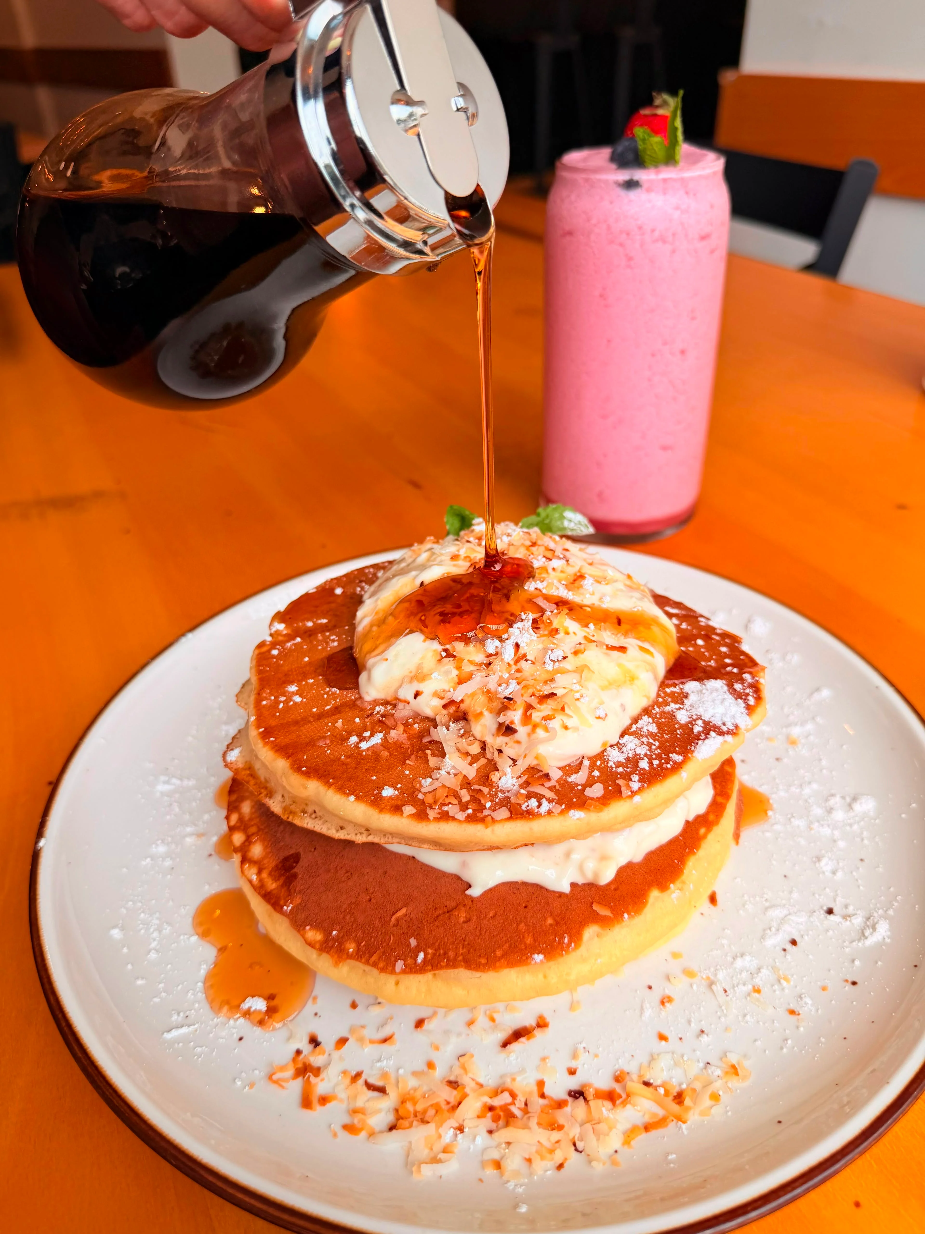 A tall stack of pancakes topped with whipped cream and shredded coconut, with syrup being poured over them, and a pink shake in the background, placed on a wooden table.