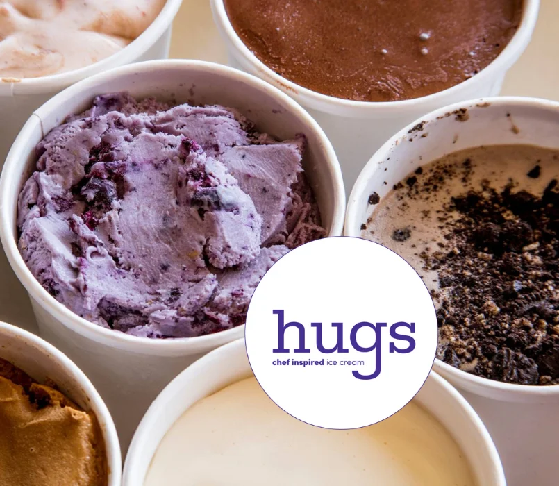 Assorted gourmet ice cream flavors in white containers from Hugs Ice Cream, featuring rich chocolate, cookies and cream, blueberry, and caramel, with the Hugs chef-inspired logo in the center.