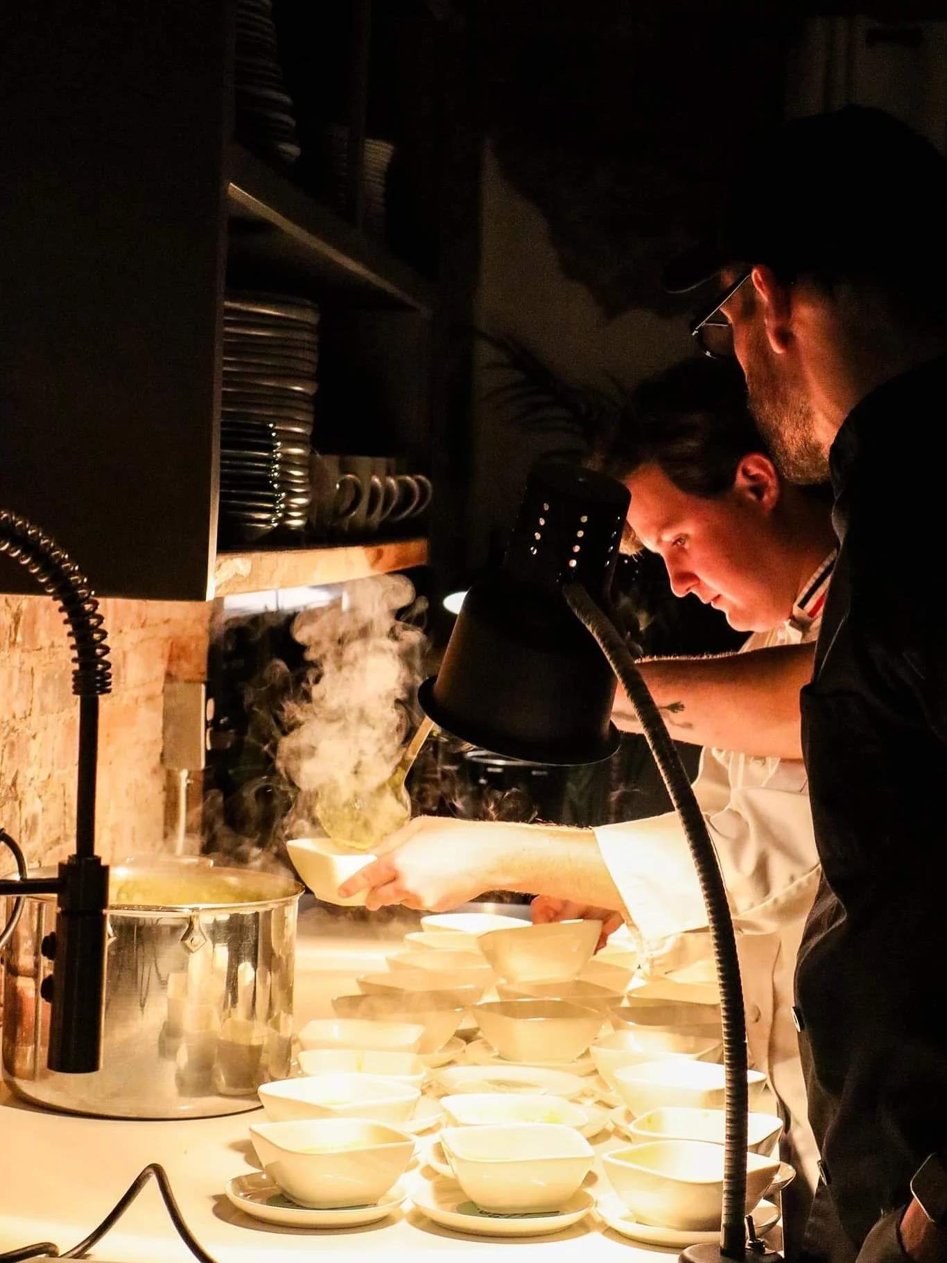 Chefs in a kitchen preparing dishes, with steam rising from pots as they serve food into bowls on a well-lit countertop.