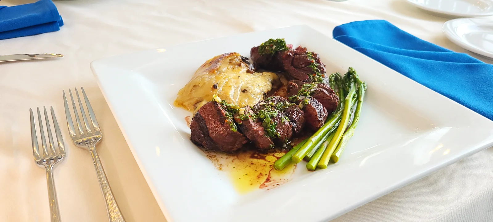 A gourmet dish featuring grilled meat slices garnished with herbs, accompanied by roasted potatoes and fresh asparagus on a square plate.