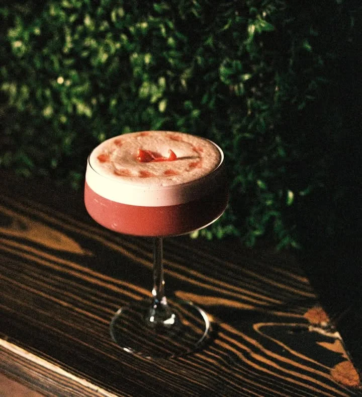 A stylish cocktail served in a coupe glass with a foamy top and garnished with small strawberry pieces.