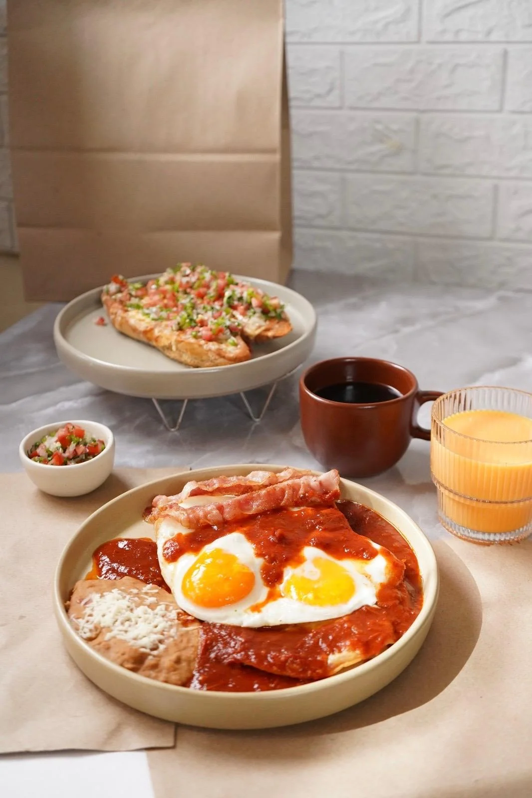 A plate of scrambled eggs topped with sausage, alongside beans and breakfast tortillas. There are beverages and a side of toast topped with fresh salsa.