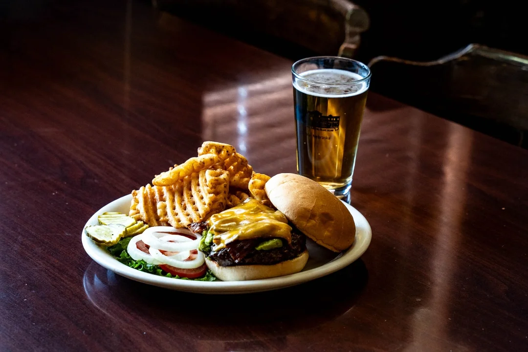 A delicious cheeseburger with melted cheese, lettuce, onions, and pickles, paired with a side of waffle fries and a beer. The meal is served on a white plate.