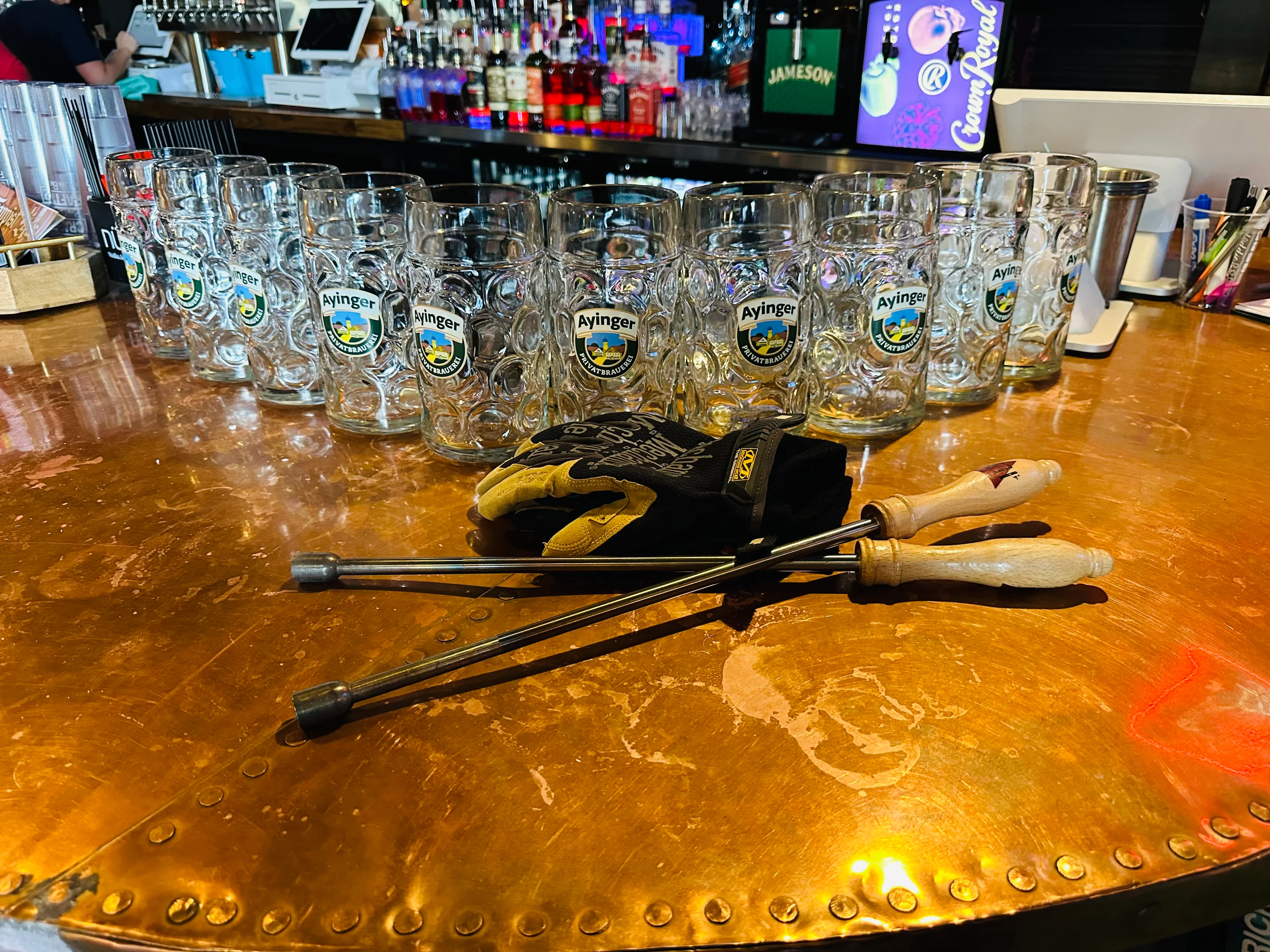 A polished bar surface with a line of empty beer glasses and a pair of tongs resting on the side. The bar has a variety of bottles visible in the background.