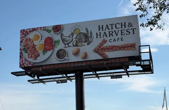 A large billboard featuring a colorful illustration of a breakfast meal with eggs, bacon, and vegetables. The restaurant name 'Hatch & Harvest Café' is prominently displayed.