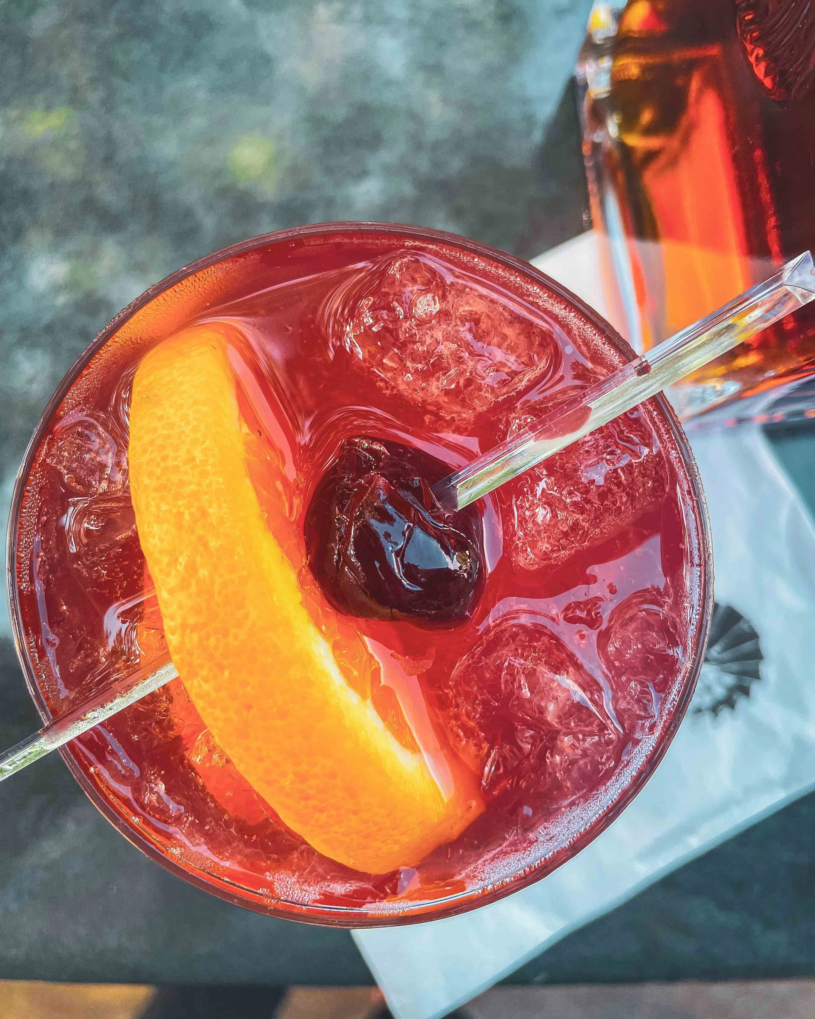 This picture shows a top-down view of a close-up of an Old Fashioned cocktail in a red color with an orange slice and cherry on top, and a napkin below the drink on the table