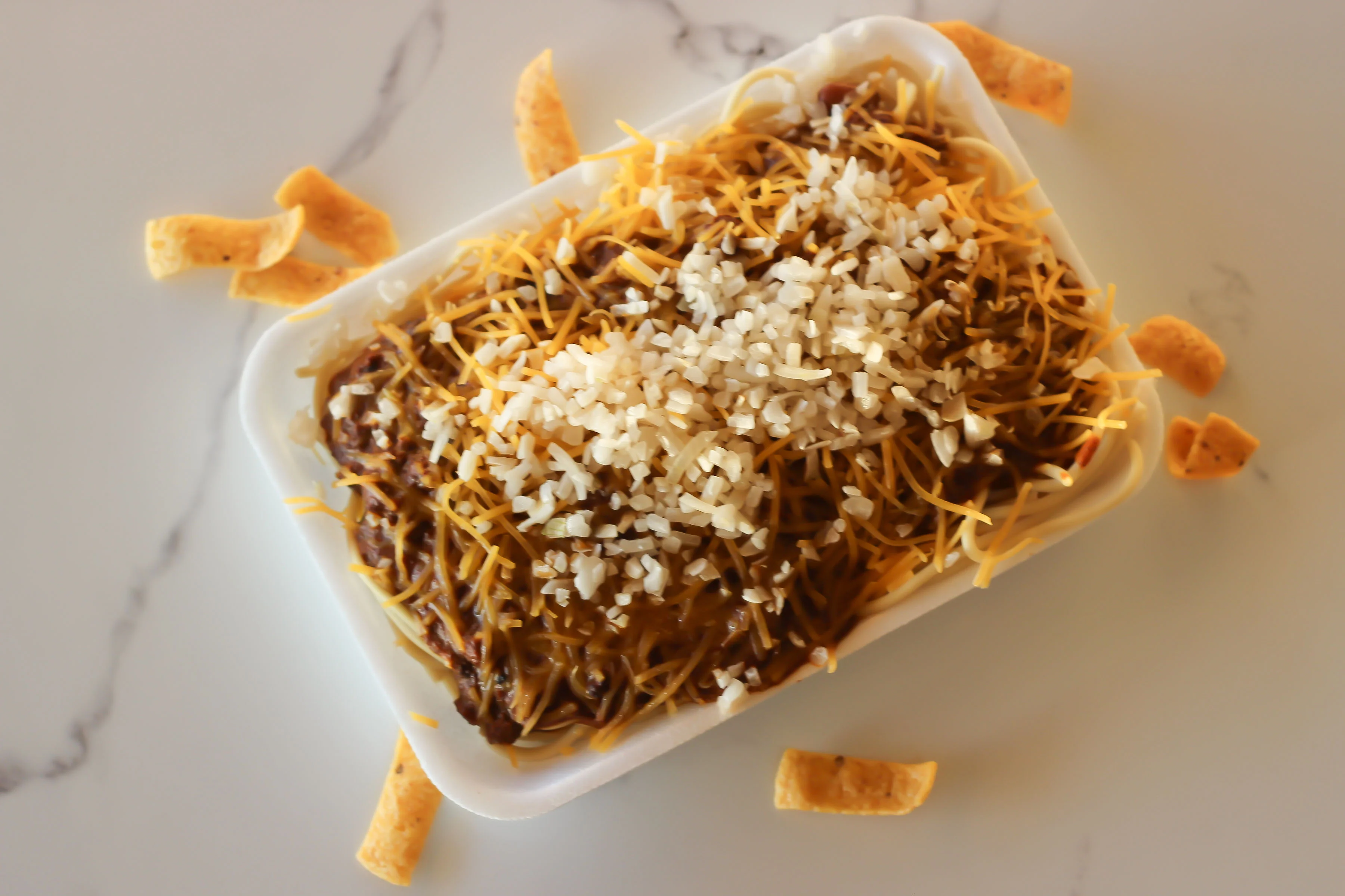 A food container filled with spaghetti topped with chili and shredded cheese, sprinkled with diced onions and surrounded by corn chips.